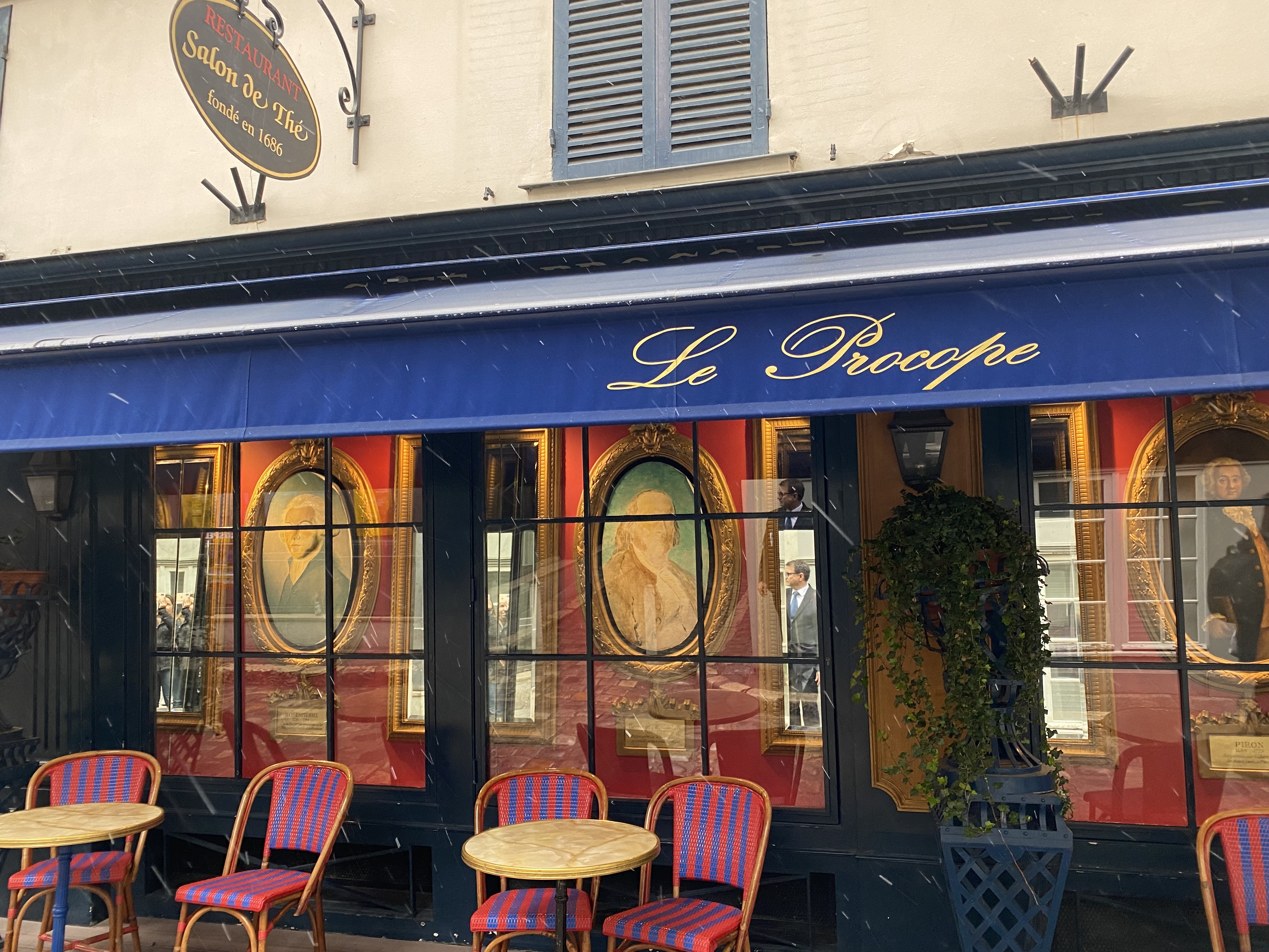 Fachada do restaurante Le Procope em Paris, com toldo azul e letras douradas. Abaixo, janelas exibem pinturas emolduradas sobre fundo vermelho. Na frente, mesas redondas e cadeiras de vime vermelhas e azuis, com neve caindo suavemente