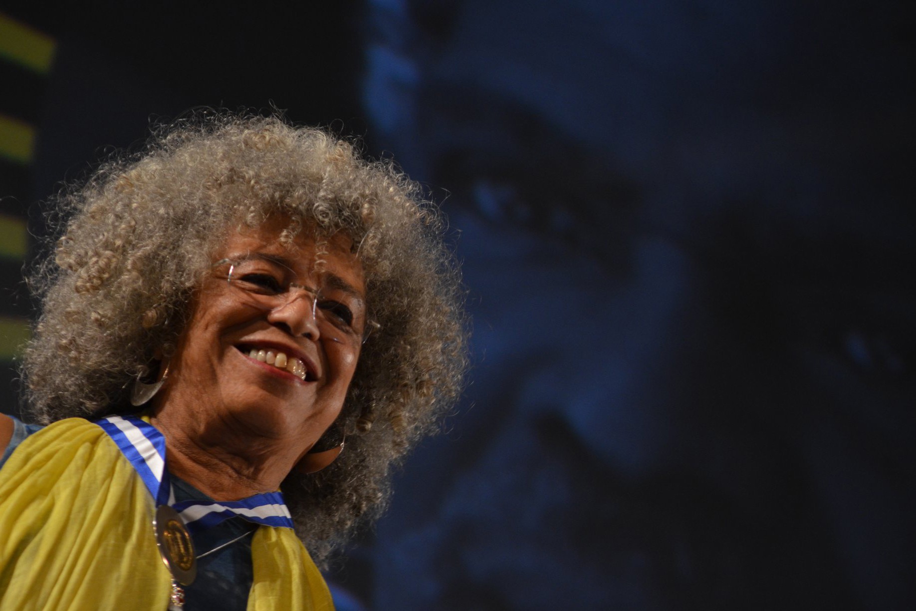 Angela Davis, mulher negra com cabelos grisalhos e crespos, sorri amplamente, usando &oacute;culos, brincos grandes e um colar com medalha sobre uma faixa azul e branca, vestindo uma blusa amarela. O fundo &eacute; escuro e desfocado