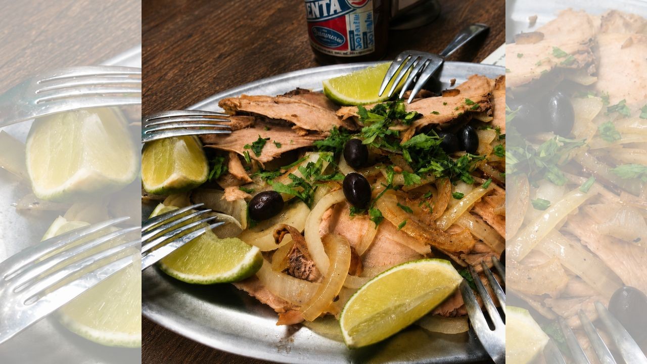 Prato de carne fatiada com cebola caramelizada, azeitonas pretas, cheiro-verde picado e fatias de limão, servido em travessa metálica com garfos. Uma garrafa de bebida aparece ao fundo