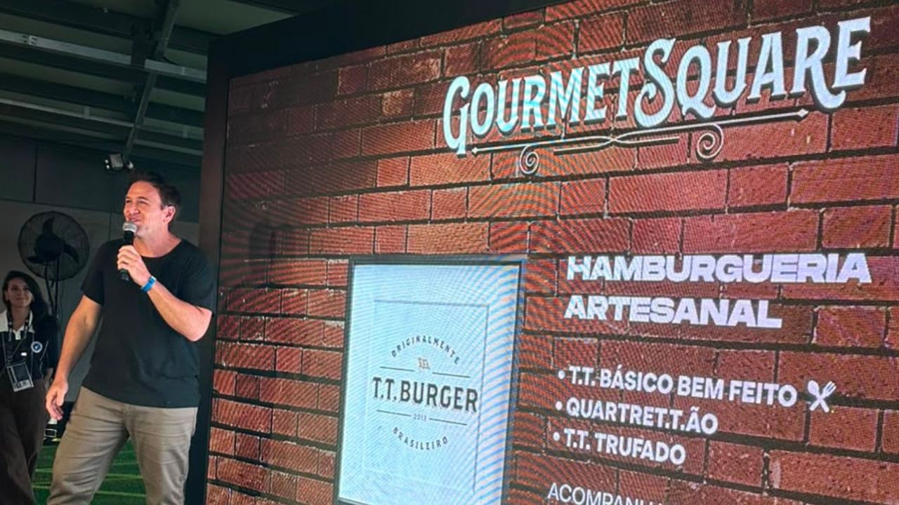 Homem sorridente de camiseta preta e calça cáqui, segurando um microfone, em frente a um telão com fundo de tijolos. No telão, lê-se GOURMET SQUARE HAMBURGUERIA ARTESANAL e o logo T.T. BURGER, com opções de hambúrgueres listadas