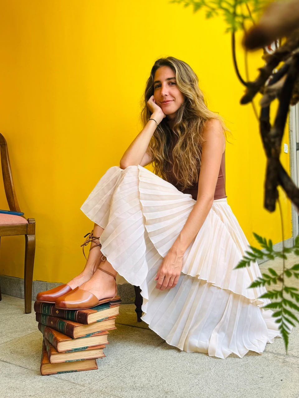 Mulher sorridente com cabelos longos e ondulados, usando blusa marrom e saia plissada bege, sentada no chão com os pés sobre uma pilha de livros antigos. O fundo é uma parede amarela vibrante, com folhagens verdes no canto superior direito e uma cadeira de madeira à esquerda