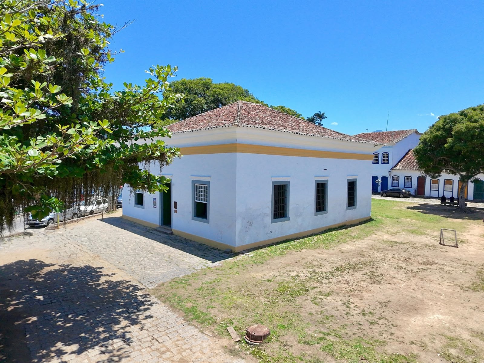 Casar&atilde;o colonial branco com telhado de barro e faixa amarela, em Paraty, RJ. &Agrave; esquerda, &aacute;rvores frondosas e rua de paralelep&iacute;pedos com carros estacionados. &Agrave; direita, um gramado e outras constru&ccedil;&otilde;es hist&oacute;ricas ao fundo, sob c&eacute;u azul claro.