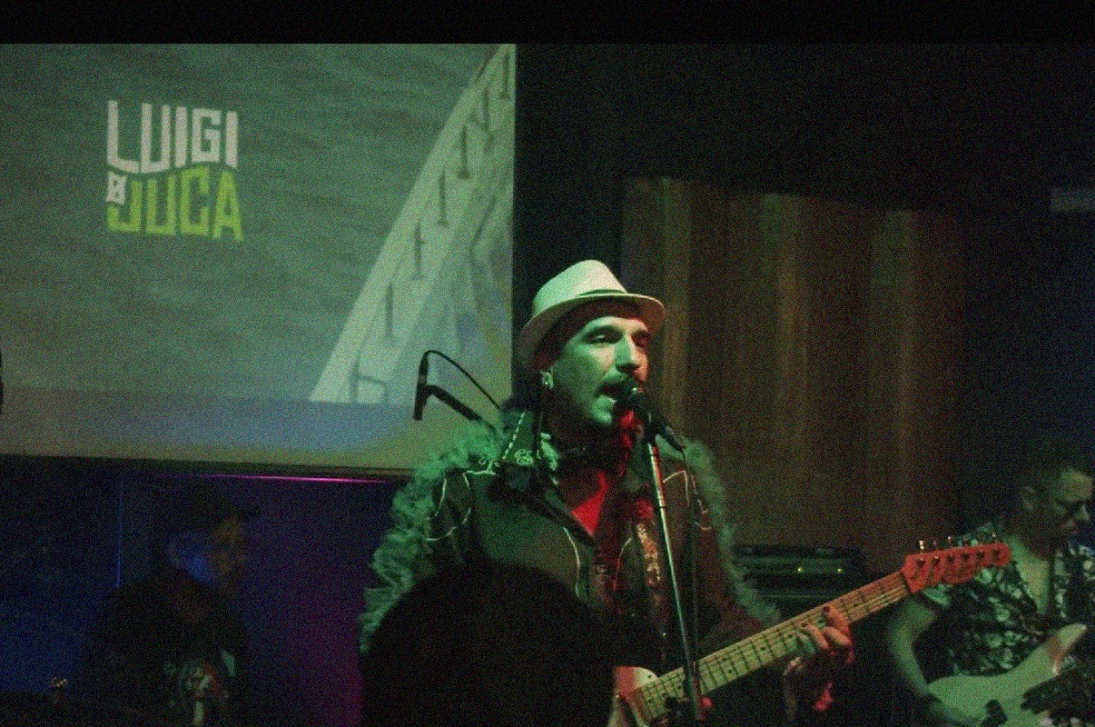 Cantor de chapéu branco e casaco peludo verde, com barba e bigode, canta no microfone e toca guitarra em palco escuro. Ao fundo, projeção verde com o nome LUIGI E LUCA. Outros músicos aparecem desfocados.