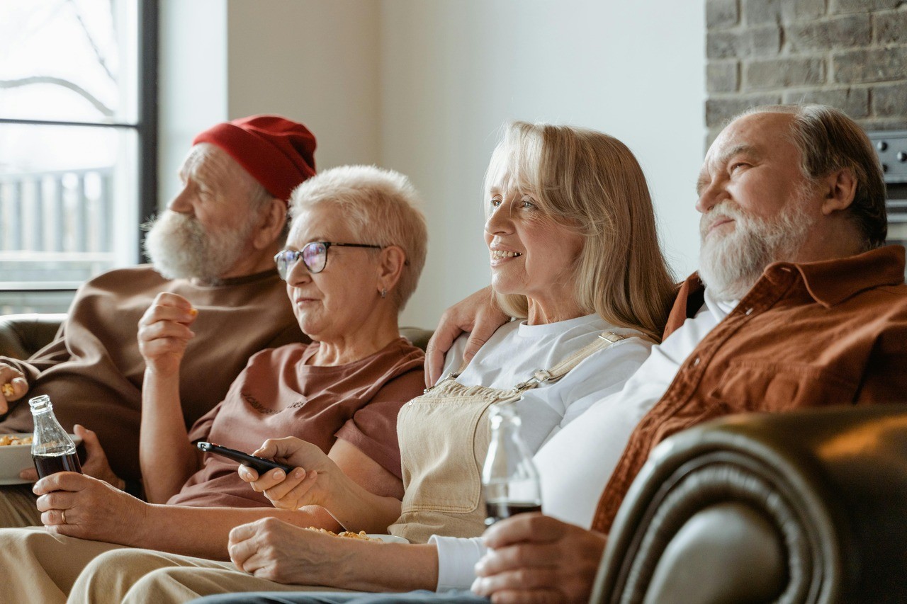 Quatro idosos, dois homens e duas mulheres, sentados no sofá assistindo TV. Um homem de gorro vermelho e barba branca come pipoca. Uma mulher de óculos segura um controle remoto. Outra mulher sorri, e um homem de camisa marrom segura uma garrafa de refrigerante. Todos parecem relaxados e desfrutando do momento juntos