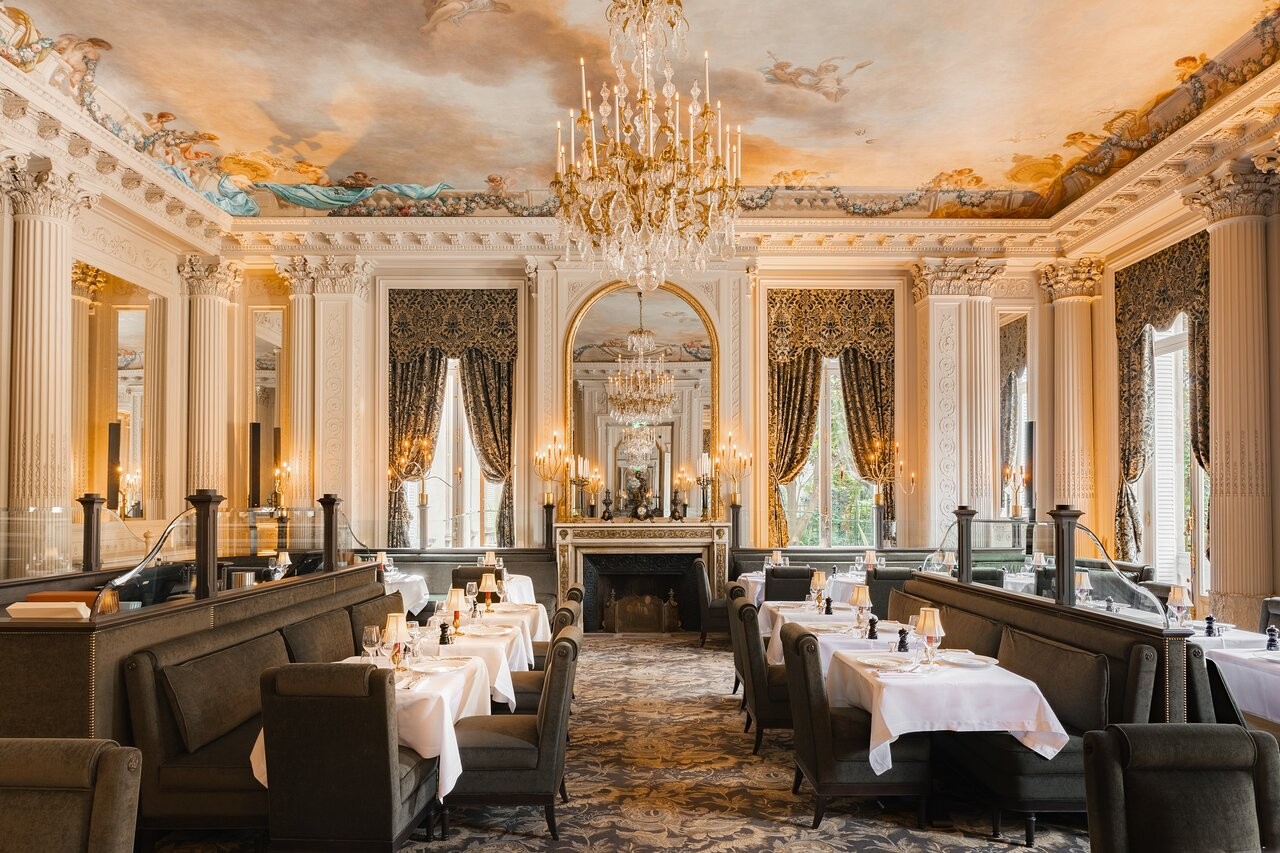 Salão de restaurante luxuoso com teto pintado de nuvens e anjos, lustres dourados, espelhos, colunas brancas e janelas com cortinas estampadas. Mesas com toalhas brancas e cadeiras estofadas em verde-oliva, dispostas em fileiras com divisórias baixas