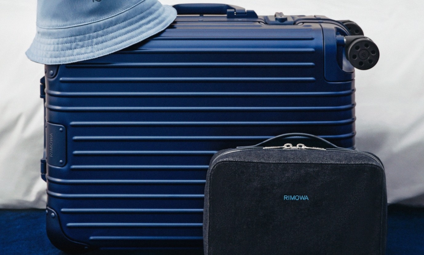 Uma mala de viagem azul-marinho com listras horizontais e um chapéu bucket azul claro sobre ela, ao lado de uma pequena bolsa preta com o nome RIMOWA em azul claro, tudo sobre um fundo branco