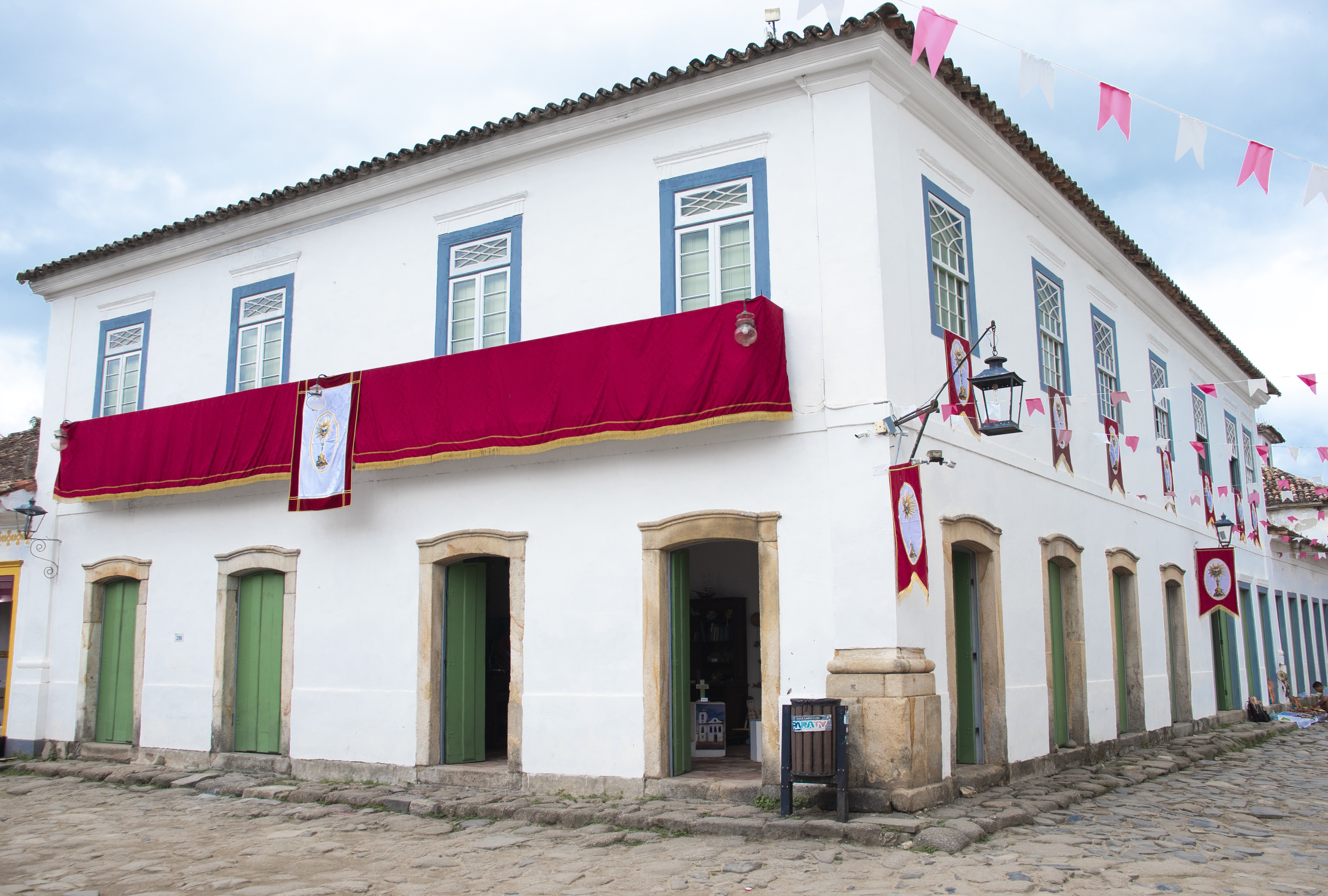 Casarão colonial branco com janelas azuis e portas verdes, decorado com um tecido vermelho no andar superior e bandeirinhas rosa e brancas penduradas no céu, em uma rua de paralelepípedos