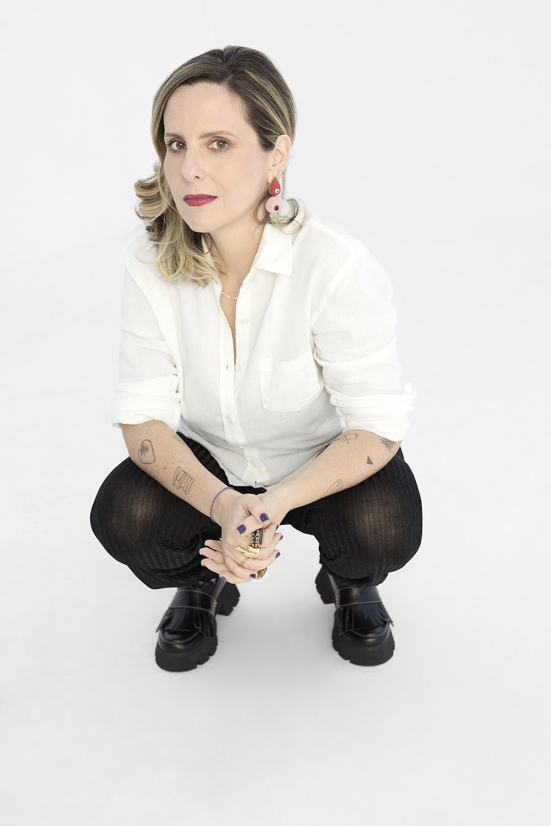 Mulher de cabelos loiros e castanhos, batom vermelho, camisa branca e calça preta, agachada com as mãos unidas sobre os joelhos. Ela usa brincos grandes, anéis dourados e sapatos pretos de plataforma, exibindo tatuagens nos braços