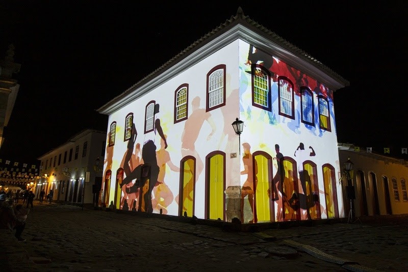 Fachada de um casarão colonial branco iluminada por projeções coloridas de silhuetas humanas dançando, com janelas e portas emolduradas em amarelo. A rua de paralelepípedos e outros prédios escuros ao fundo criam um ambiente noturno festivo