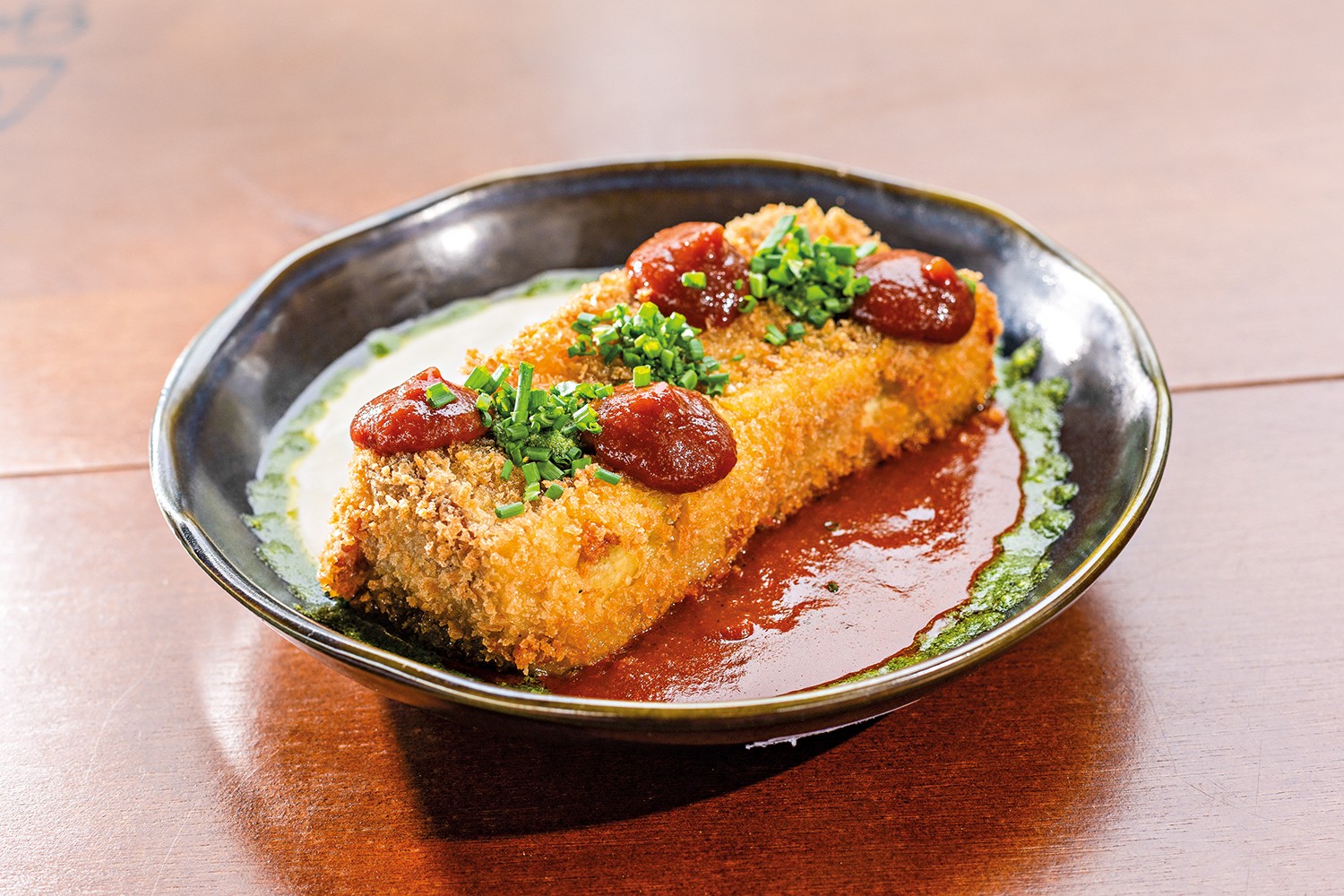 Um prato com um bolinho retangular empanado e frito, coberto por molho vermelho e cebolinha picada, servido sobre molho branco e um fio de molho verde, em uma mesa de madeira