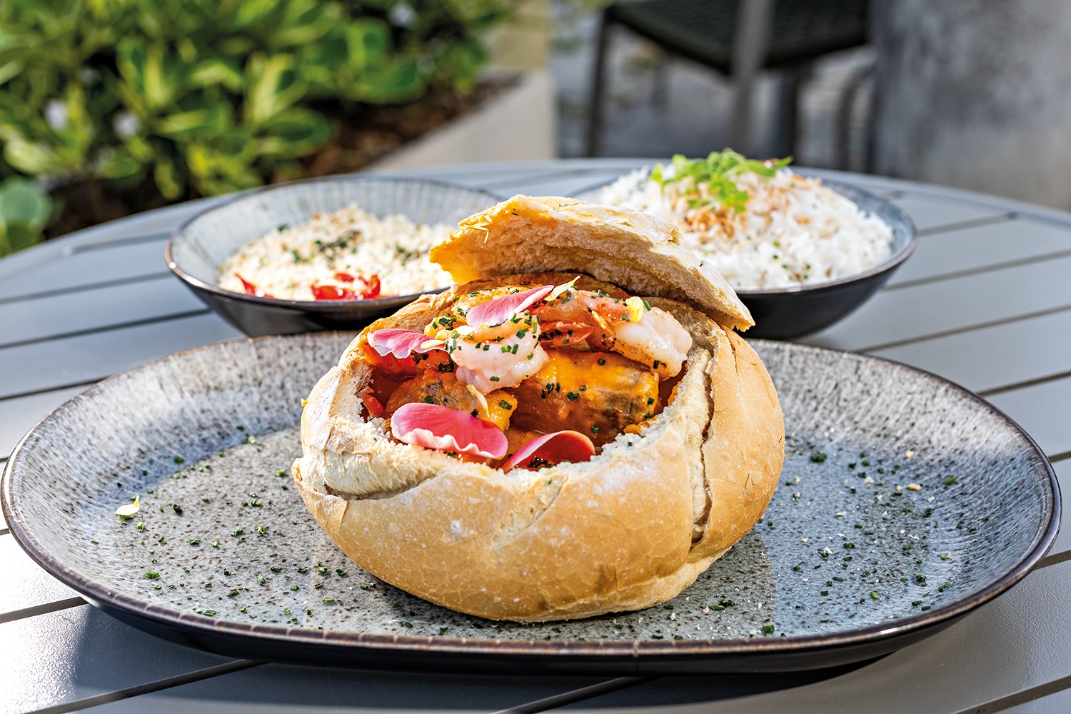 Um pão italiano redondo recheado com moqueca de camarão, decorado com pétalas de flores comestíveis e cebolinha picada, servido em um prato cinza texturizado. Ao fundo, duas tigelas com arroz branco e temperos, sobre uma mesa de madeira clara, com plantas verdes desfocadas