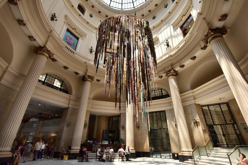 Interior de um edifício grandioso com teto abobadado e claraboia de vidro, colunas coríntias e varandas em vários níveis. Um lustre circular enorme, feito de tiras de tecido multicoloridas penduradas, domina o centro. Pessoas pequenas circulam no térreo, algumas sentadas em mesas, outras caminhando. A luz do sol entra pelas janelas e claraboia, iluminando o espaço.