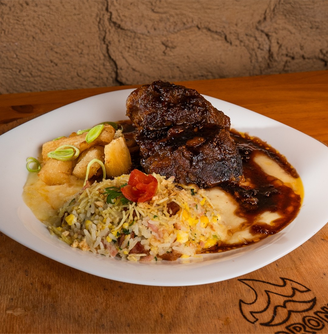 Prato com carne escura em molho, purê cremoso, mandioca frita com cebolinha e arroz colorido com bacon e ovo, decorado com tomate cereja