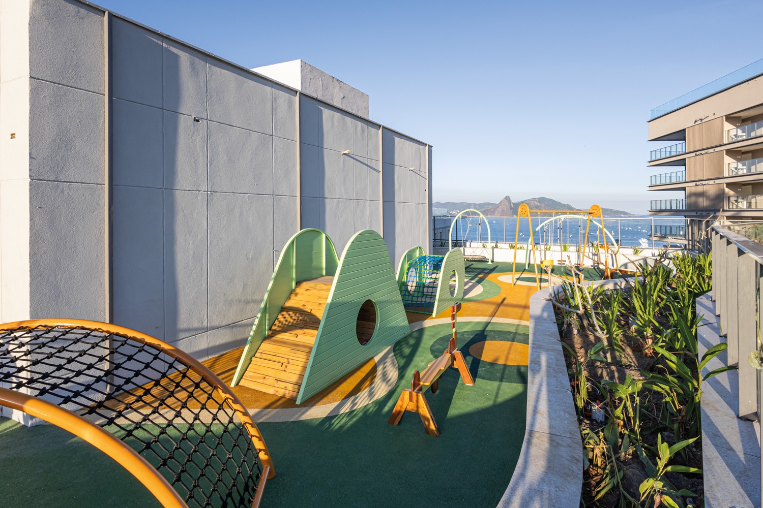 Parque infantil em um terra&ccedil;o com vista para o mar e o P&atilde;o de A&ccedil;&uacute;car, com brinquedos coloridos em tons de verde, amarelo e azul, e um edif&iacute;cio moderno &agrave; direita