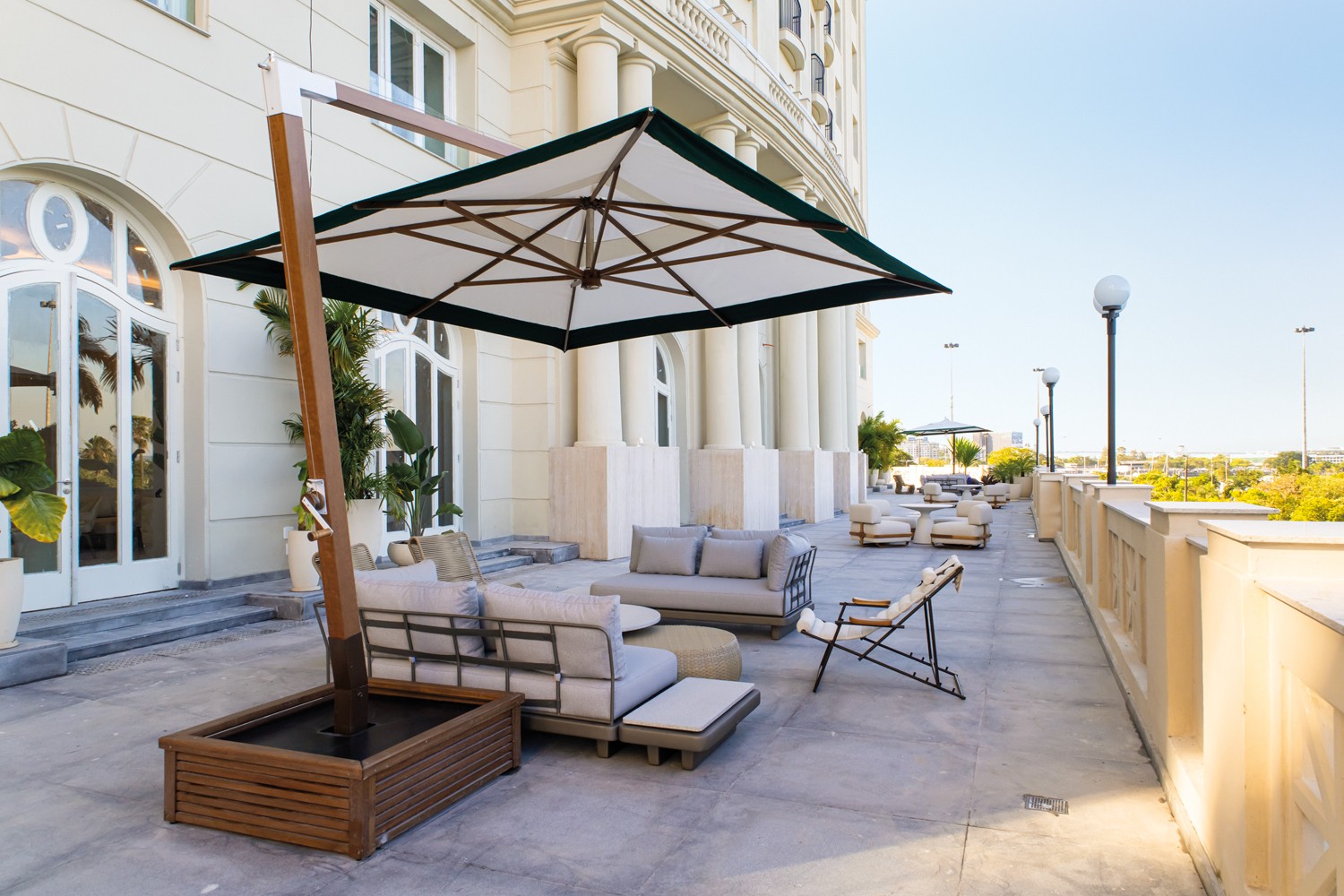 Terra&ccedil;o de hotel luxuoso com piso de cimento, mobili&aacute;rio de exterior cinza e bege, e um grande guarda-sol quadrado branco e verde. Ao fundo, um edif&iacute;cio cl&aacute;ssico com colunas e janelas arqueadas, e um c&eacute;u azul claro.