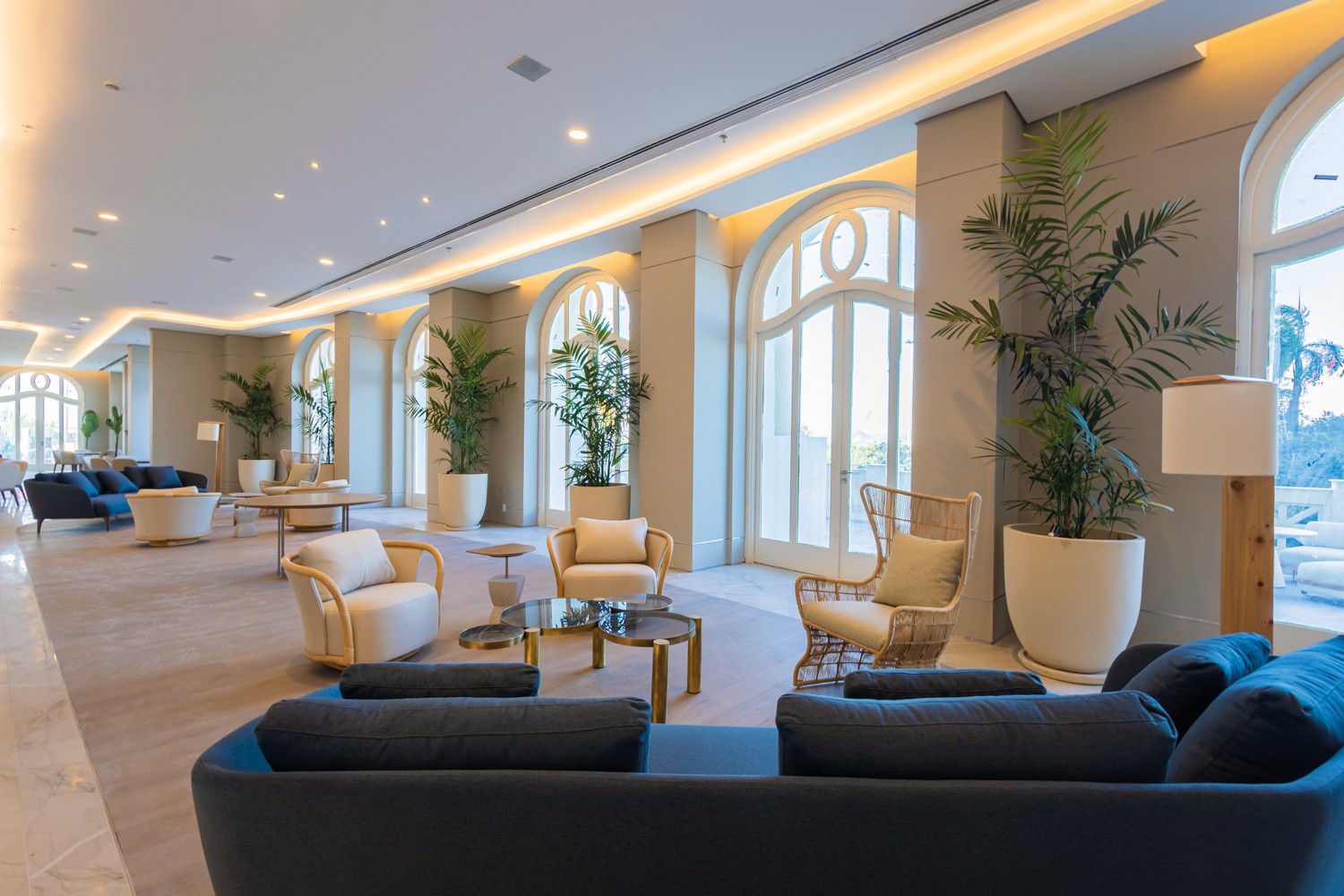 Lounge amplo e iluminado com sof&aacute;s azuis, poltronas claras e mesas de centro. Grandes janelas arqueadas e plantas altas em vasos brancos decoram o ambiente