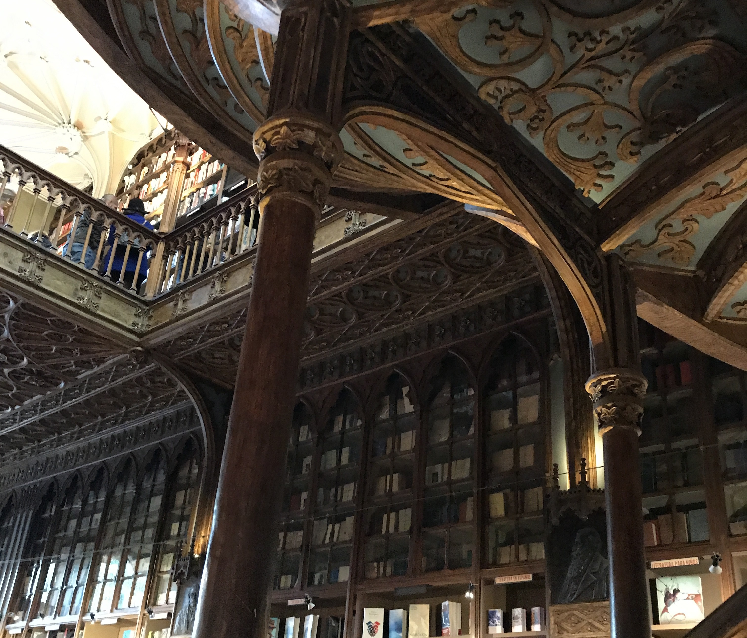 Interior de uma livraria antiga com arquitetura gótica. Prateleiras de madeira escura com livros preenchem as paredes. Um balcão superior com pessoas e grades ornamentadas é visível. Colunas e arcos de madeira entalhada sustentam um teto azul claro com detalhes dourados. A iluminação é suave, criando uma atmosfera acolhedora e histórica