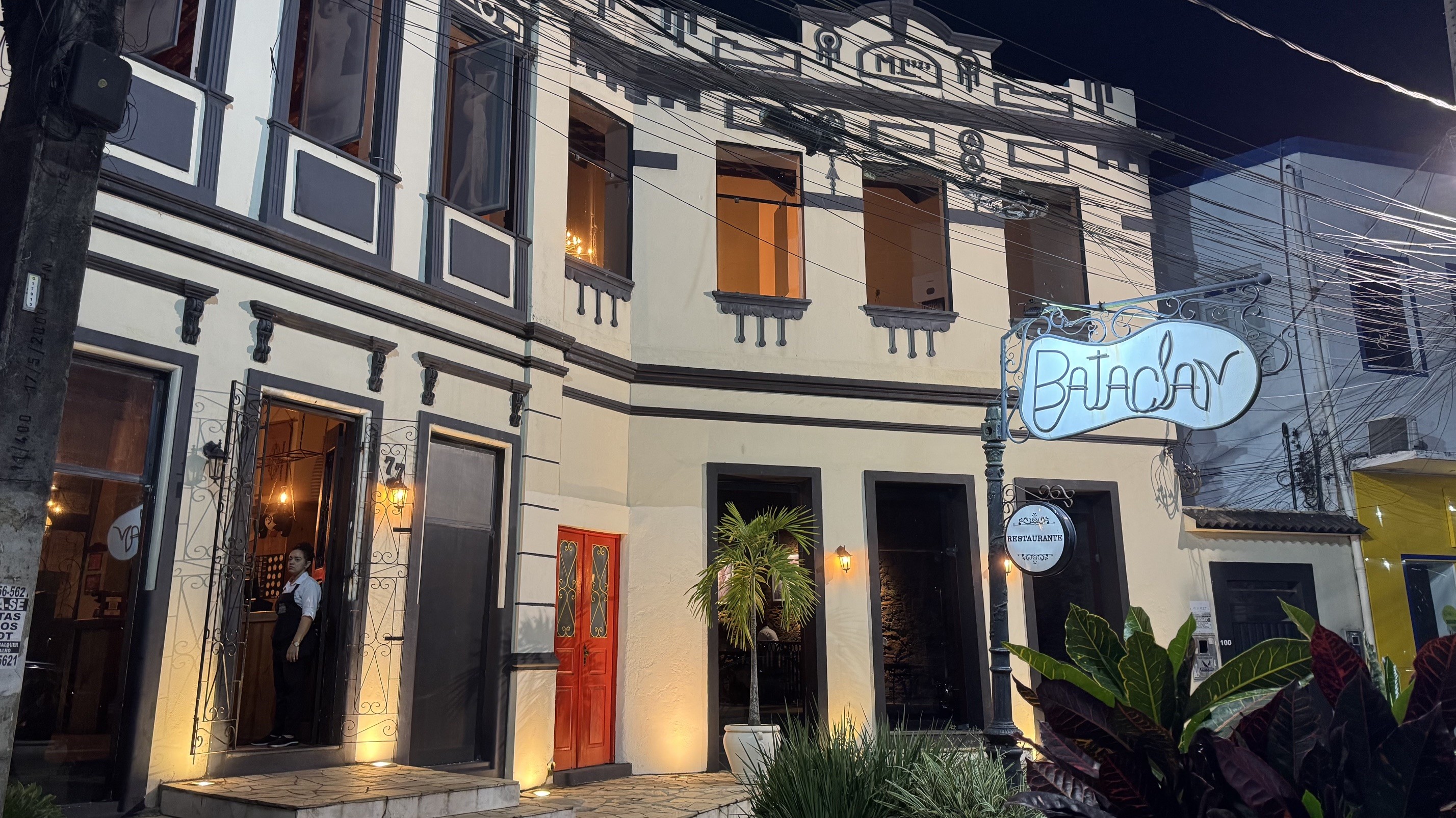 Fachada noturna de um prédio de dois andares, bege com detalhes em cinza escuro. Um letreiro luminoso em formato de violão com a palavra Bataclan está pendurado na frente. Um homem de camisa branca e avental escuro aparece na porta de entrada. Há plantas verdes em vasos e uma porta vermelha ao lado. Fios elétricos cruzam a imagem no alto