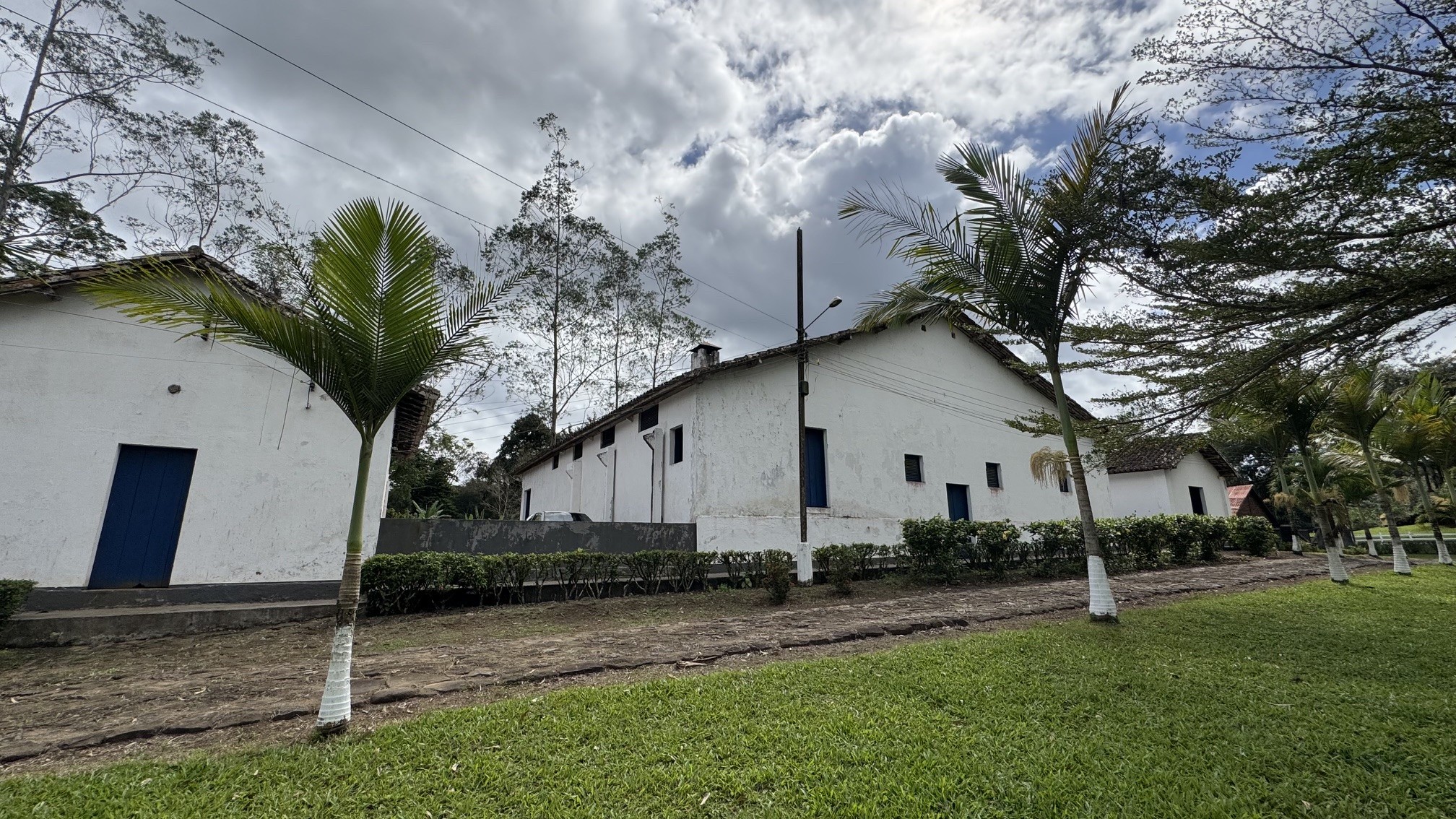 Três casas brancas com telhados de barro e portas azuis, cercadas por gramado verde, terra batida e palmeiras, sob céu nublado