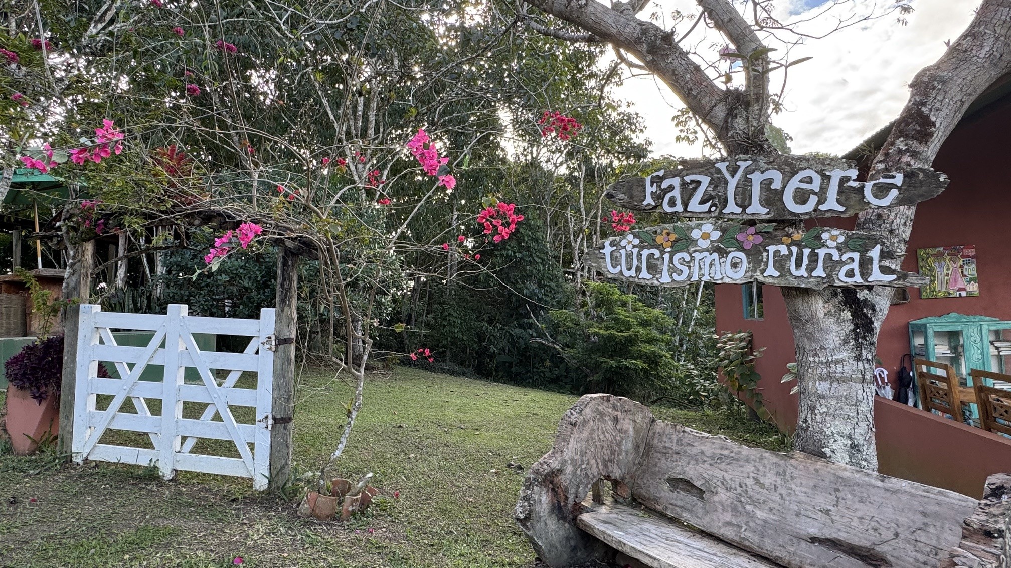 Um portão branco de madeira em um jardim verde com árvores e flores cor-de-rosa. À direita, um banco rústico de madeira e uma placa indicando FazYrere turismo rural em um tronco de árvore. Ao fundo, uma casa com paredes avermelhadas e janelas. O céu está parcialmente nublado