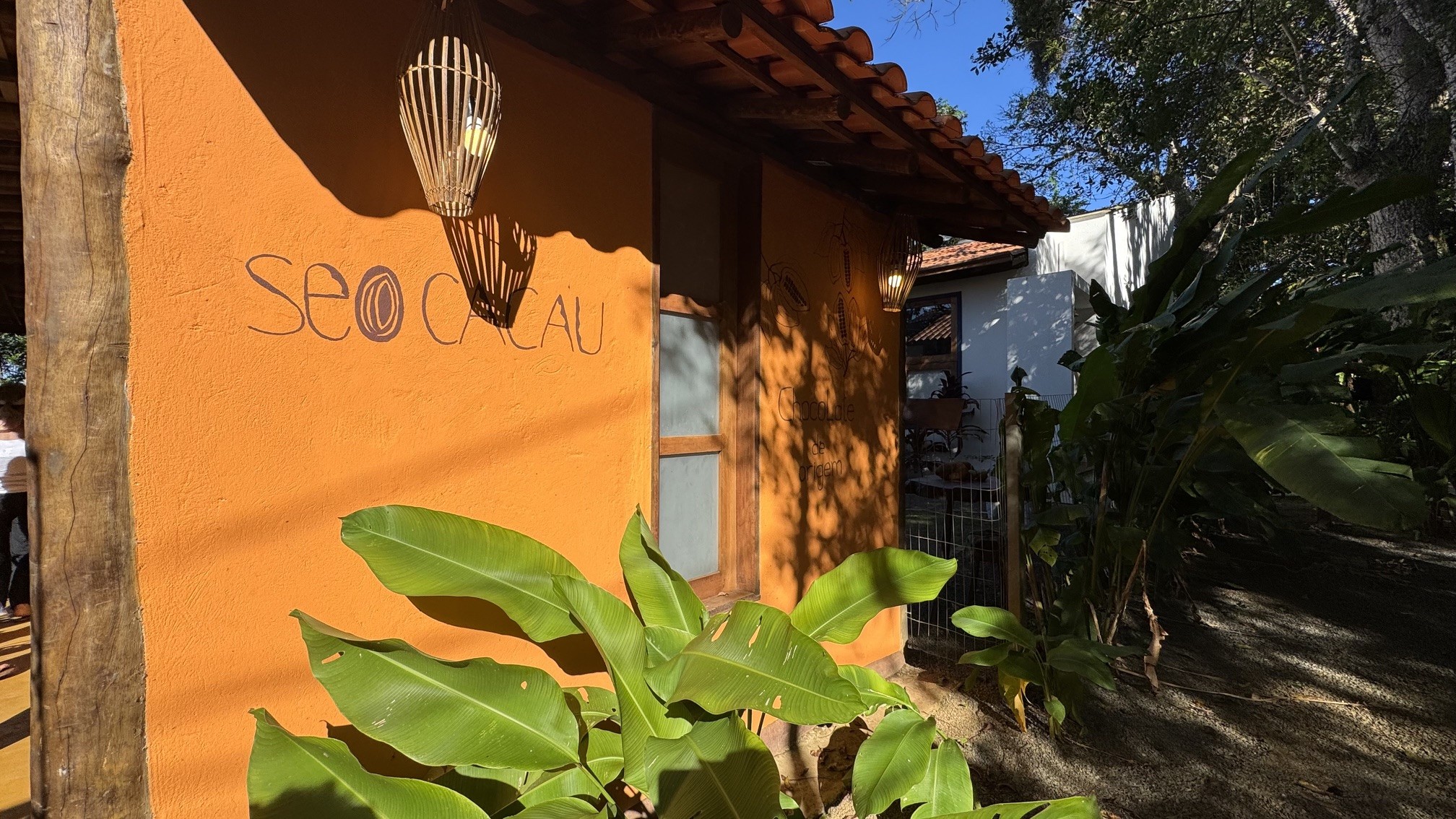 Fachada de um estabelecimento com parede laranja-terra, telhado de telhas cerâmicas e o nome Seo Cacau escrito em preto. Há um poste de madeira à esquerda, uma janela de madeira e vidro, e duas luminárias de bambu penduradas. Plantas verdes grandes em primeiro plano e vegetação densa ao fundo, sob céu azul