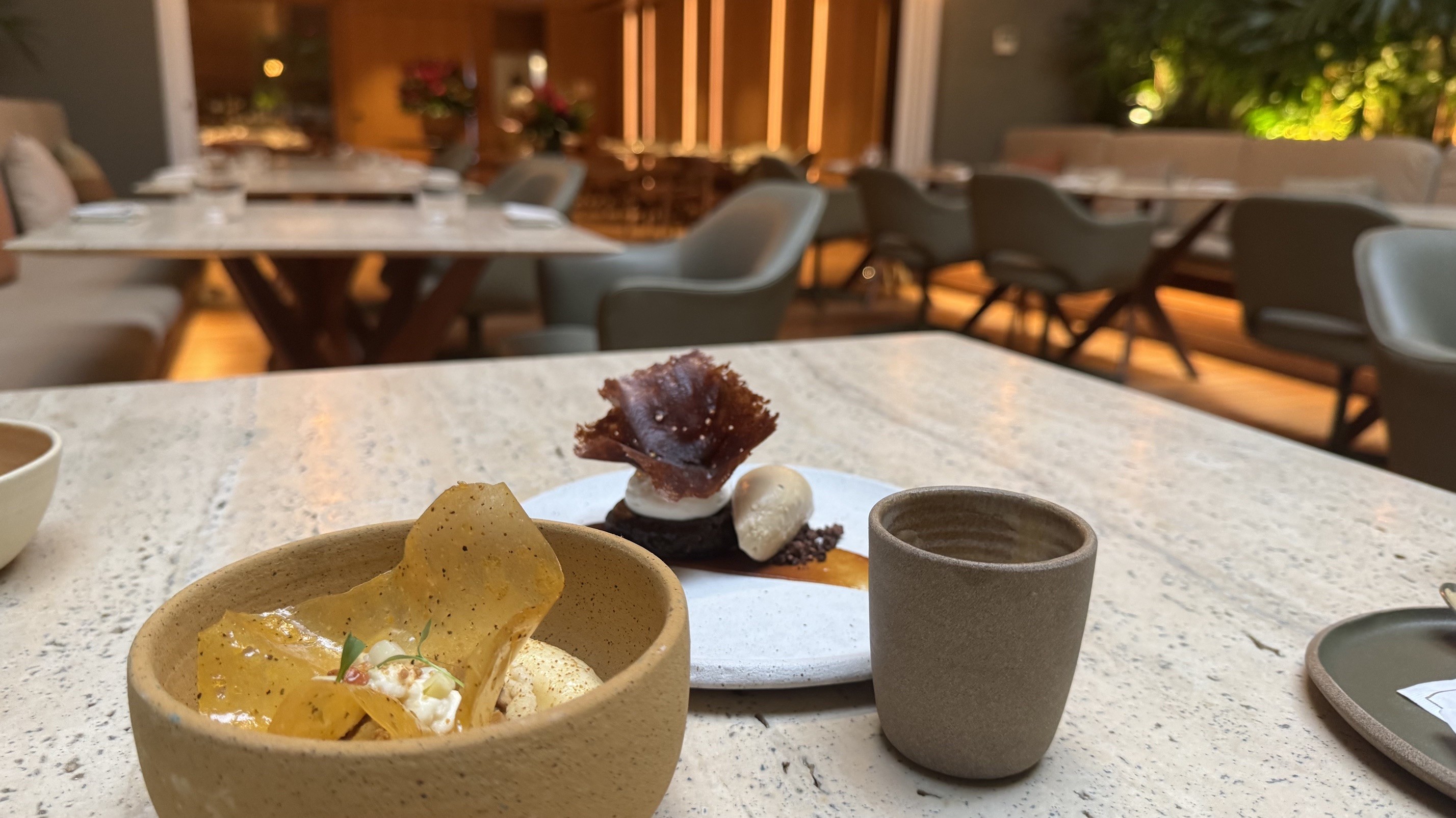 Dois pratos de sobremesa em uma mesa de pedra clara, com um copo marrom ao lado. Ao fundo, um restaurante elegante com mesas e cadeiras vazias, iluminação suave e plantas
