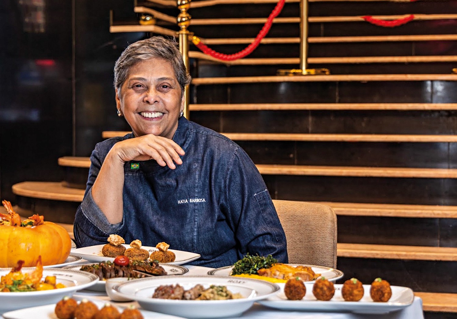 Mulher de cabelos grisalhos e sorriso largo, vestindo um dólmã jeans com o nome Katia Barbosa, sentada à mesa com diversos pratos de comida brasileira, como camarão na moranga e bolinhos, em um restaurante com escada ao fundo