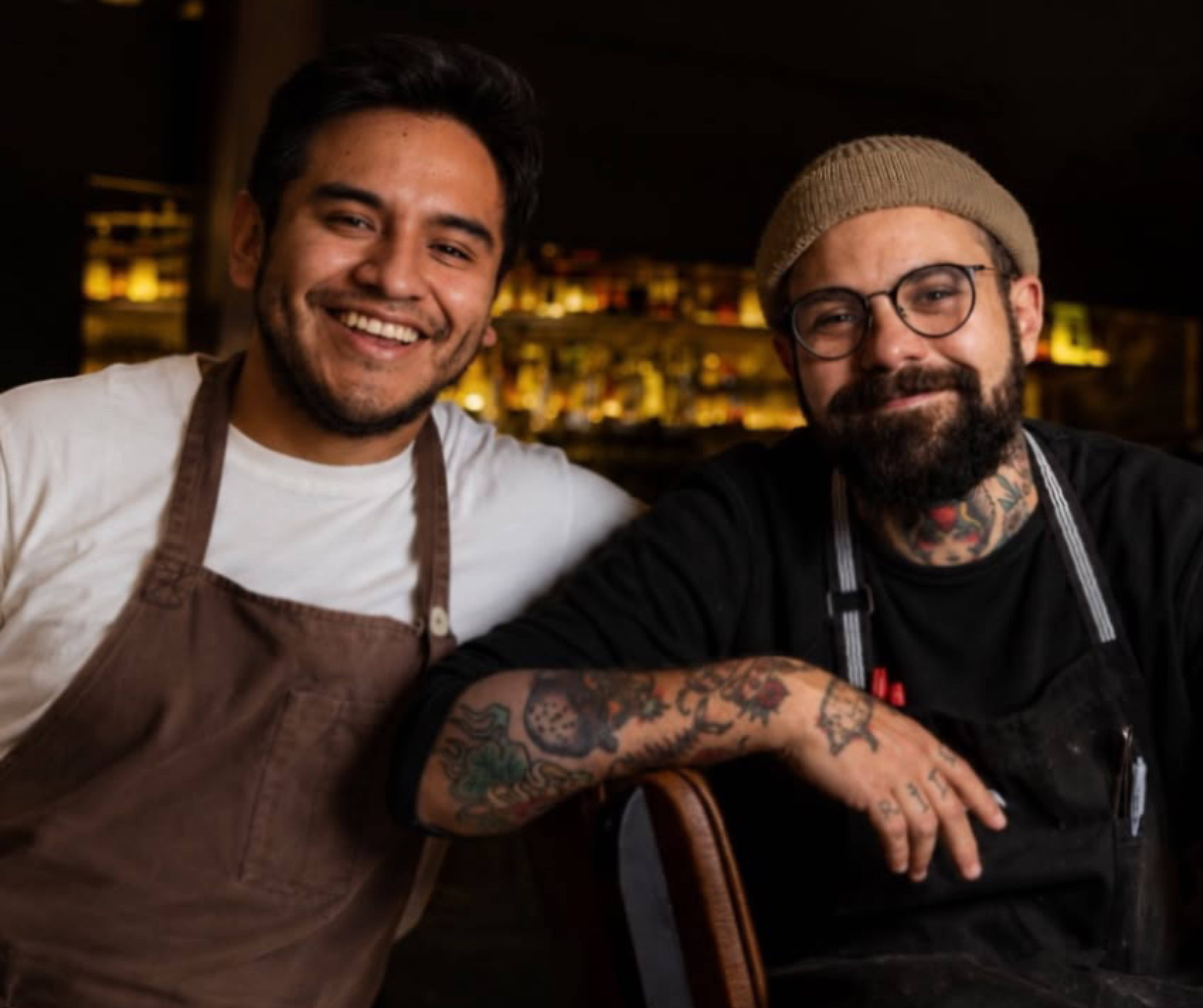 Dois homens sorridentes, um com avental marrom e outro com gorro bege, óculos e braços tatuados, posam em ambiente escuro com luzes ao fundo
