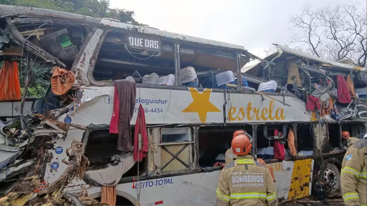 Ônibus branco da empresa Estrela, com uma estrela amarela, completamente destruído após acidente, com janelas quebradas e partes amassadas. Roupas e pertences pendurados, e dois bombeiros com capacetes laranja e uniformes bege observam a cena, com árvores secas ao fundo