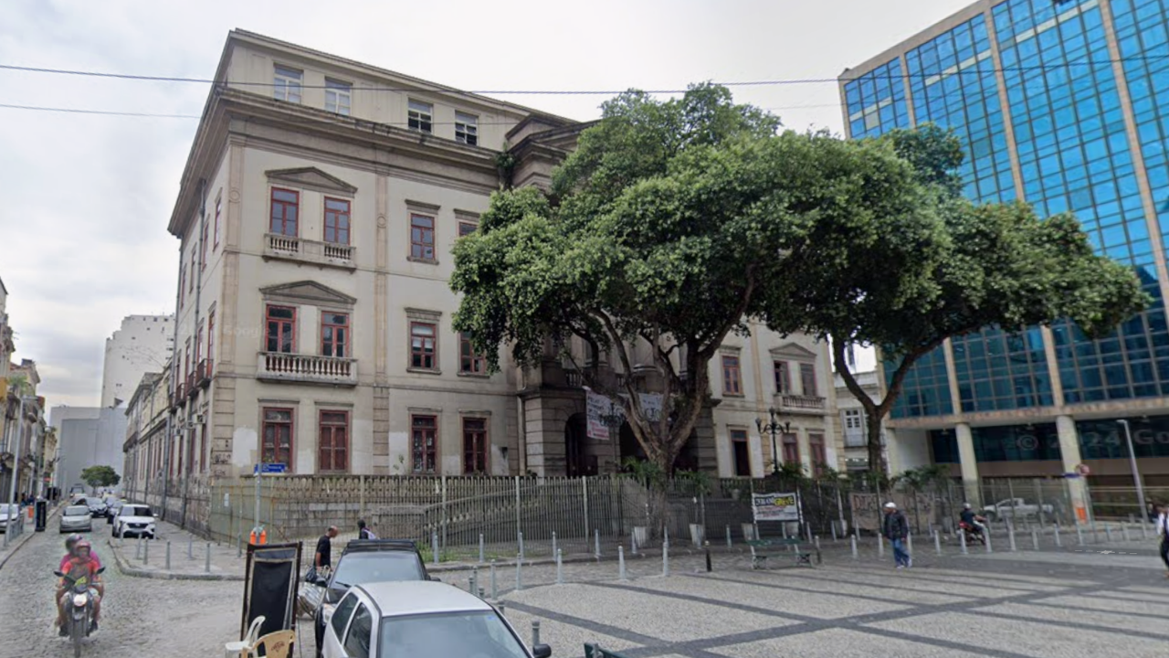 Edifício histórico de três andares com fachada bege e janelas marrons, ladeado por uma árvore frondosa. À direita, um prédio moderno de vidro azul. Em primeiro plano, uma rua de paralelepípedos com carros estacionados e pessoas caminhando. Um motociclista passa à esquerda. O céu está nublado