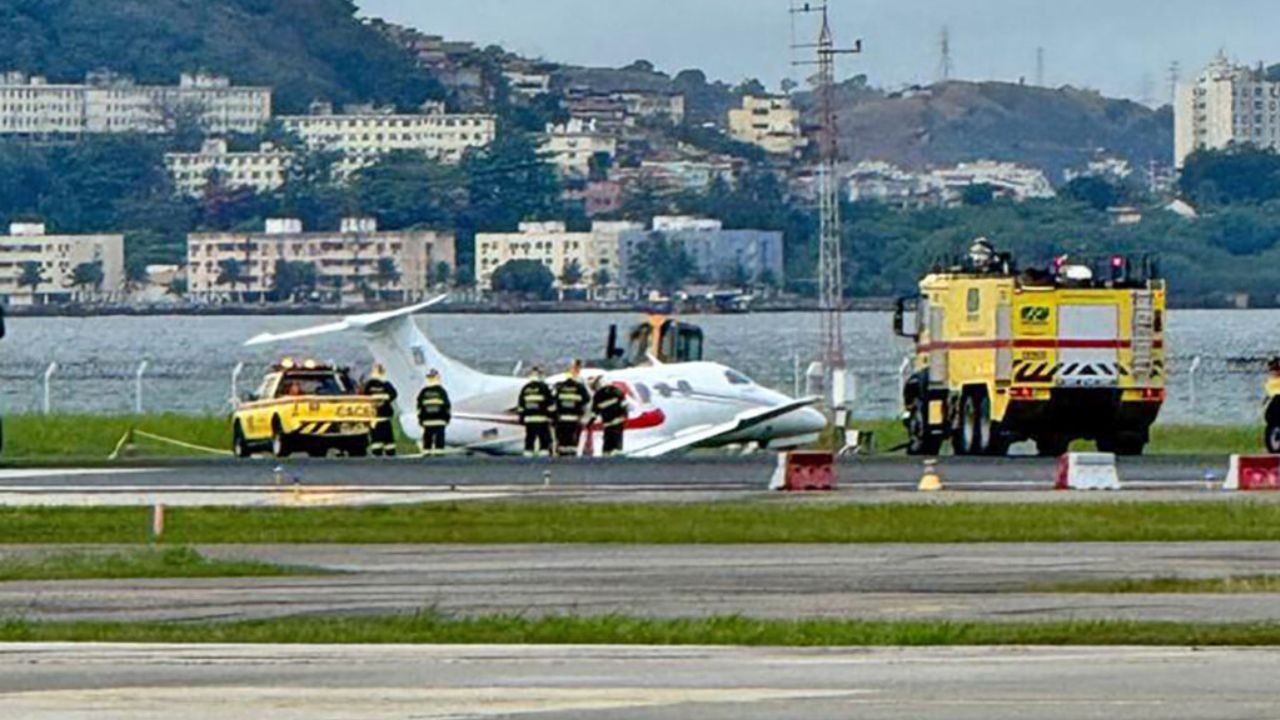 Avião branco com faixa vermelha na lateral, com o nariz no chão, cercado por bombeiros e veículos de resgate amarelos em uma pista de aeroporto, com o mar e prédios ao fundo