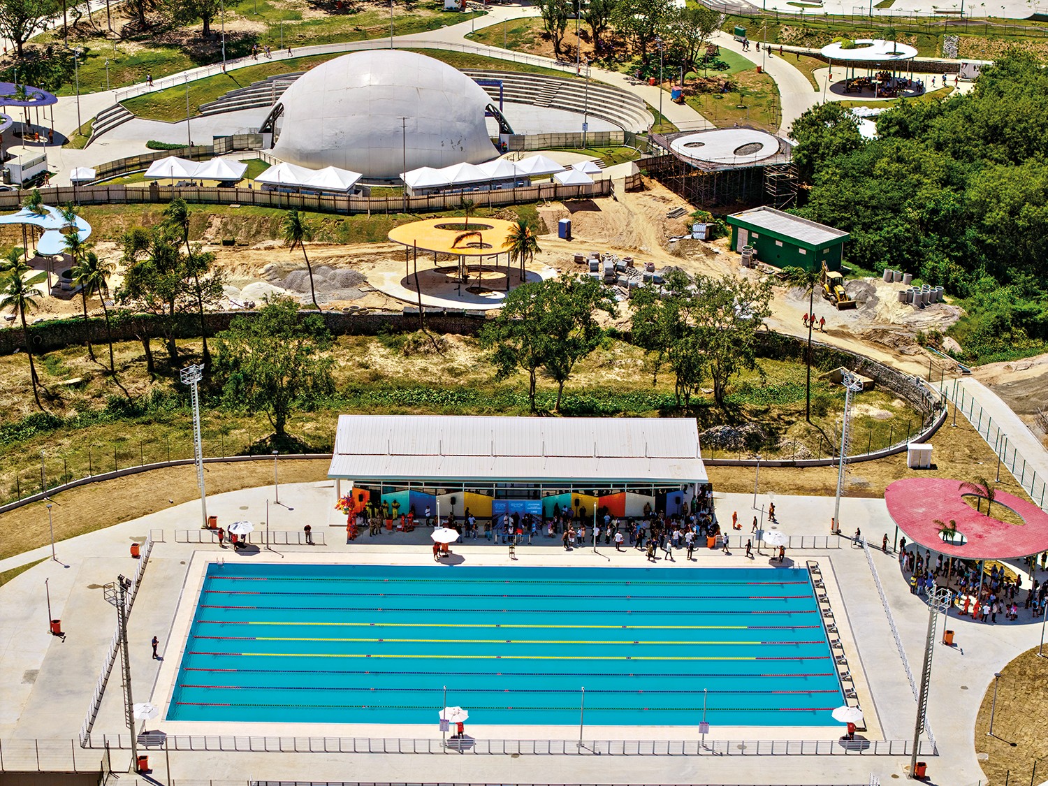 Vista a&eacute;rea de um complexo esportivo com uma piscina ol&iacute;mpica azul, cercada por pessoas e um edif&iacute;cio branco com pain&eacute;is coloridos. Ao fundo, uma grande c&uacute;pula branca e outras estruturas em constru&ccedil;&atilde;o, com &aacute;reas verdes e &aacute;rvores. O c&eacute;u est&aacute; claro e ensolarado