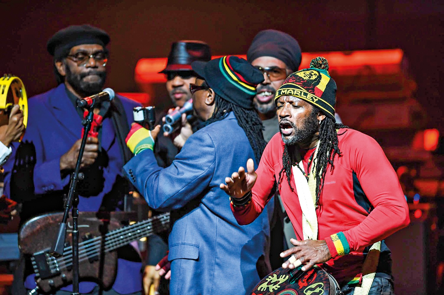 Um grupo de m&uacute;sicos negros em um palco escuro. &Agrave; esquerda, um homem de &oacute;culos e boina preta toca baixo. No centro, outro homem de chap&eacute;u e &oacute;culos escuros segura um microfone. &Agrave; direita, um cantor com dreadlocks e gorro Marley vermelho, amarelo e verde, canta com a boca aberta, batucando um tambor. Todos usam roupas escuras, exceto o cantor da direita, que veste uma camisa vermelha