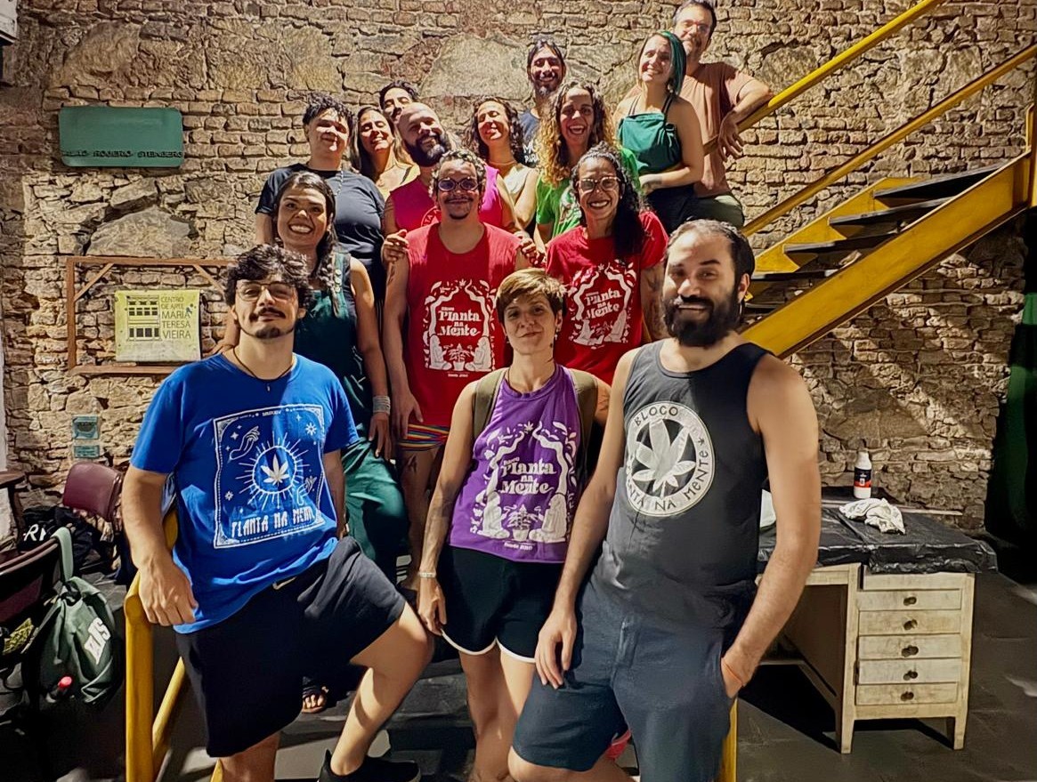 Grupo de pessoas sorrindo em frente a uma parede de tijolos e uma escada amarela. Muitos vestem camisetas com a frase Planta na Mente e um desenho de folha de cannabis.