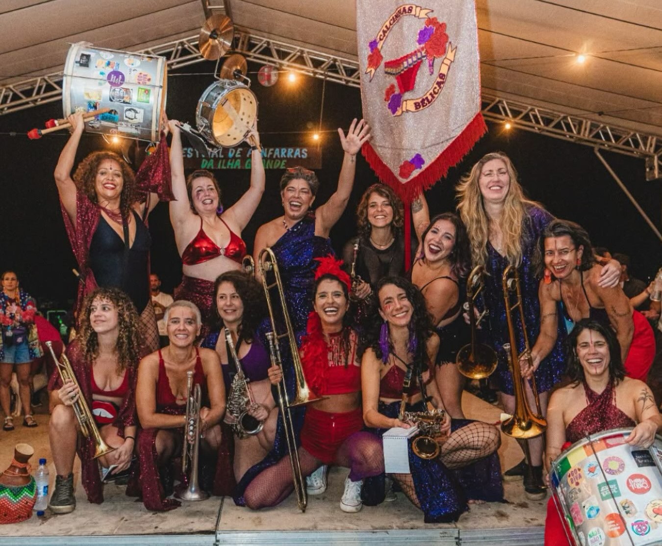 Grupo de mulheres sorrindo e posando com instrumentos musicais em um palco, algumas segurando tambores e trombones. Elas vestem roupas coloridas e brilhantes, típicas de carnaval. Ao fundo, uma faixa com o nome CALCINHAS BELICAS e luzes penduradas. O ambiente é festivo e noturno