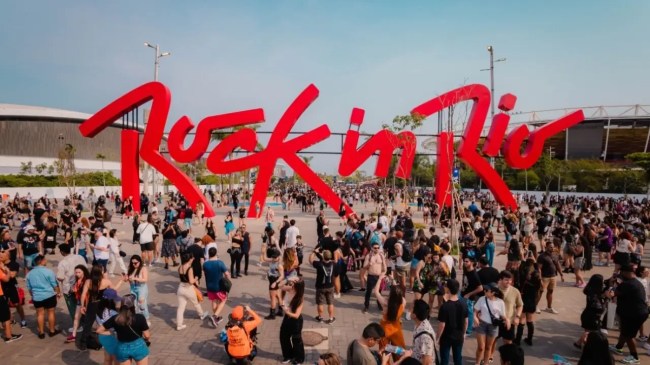 Multidão de pessoas diversas, vestindo roupas casuais, caminhando em direção ao grande letreiro vermelho Rock in Rio em um dia ensolarado. Ao fundo, estruturas de um estádio e árvores.