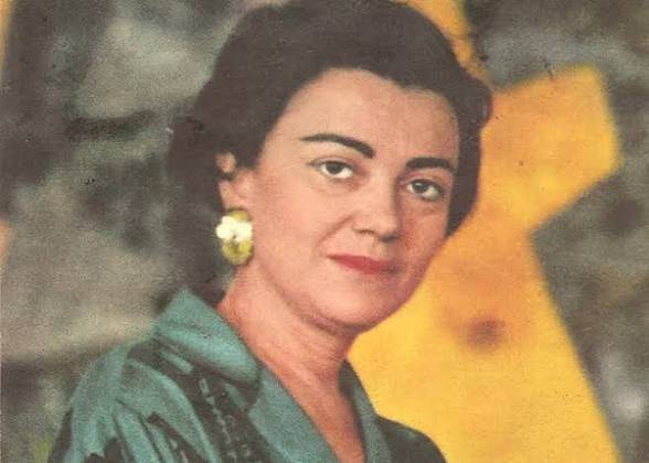 Mulher de pele clara, cabelos escuros e curtos, com batom vermelho e brincos grandes em formato de flor amarela, vestindo uma blusa verde-água, posa sorrindo levemente para a câmera, com um fundo abstrato em tons de amarelo e cinza
