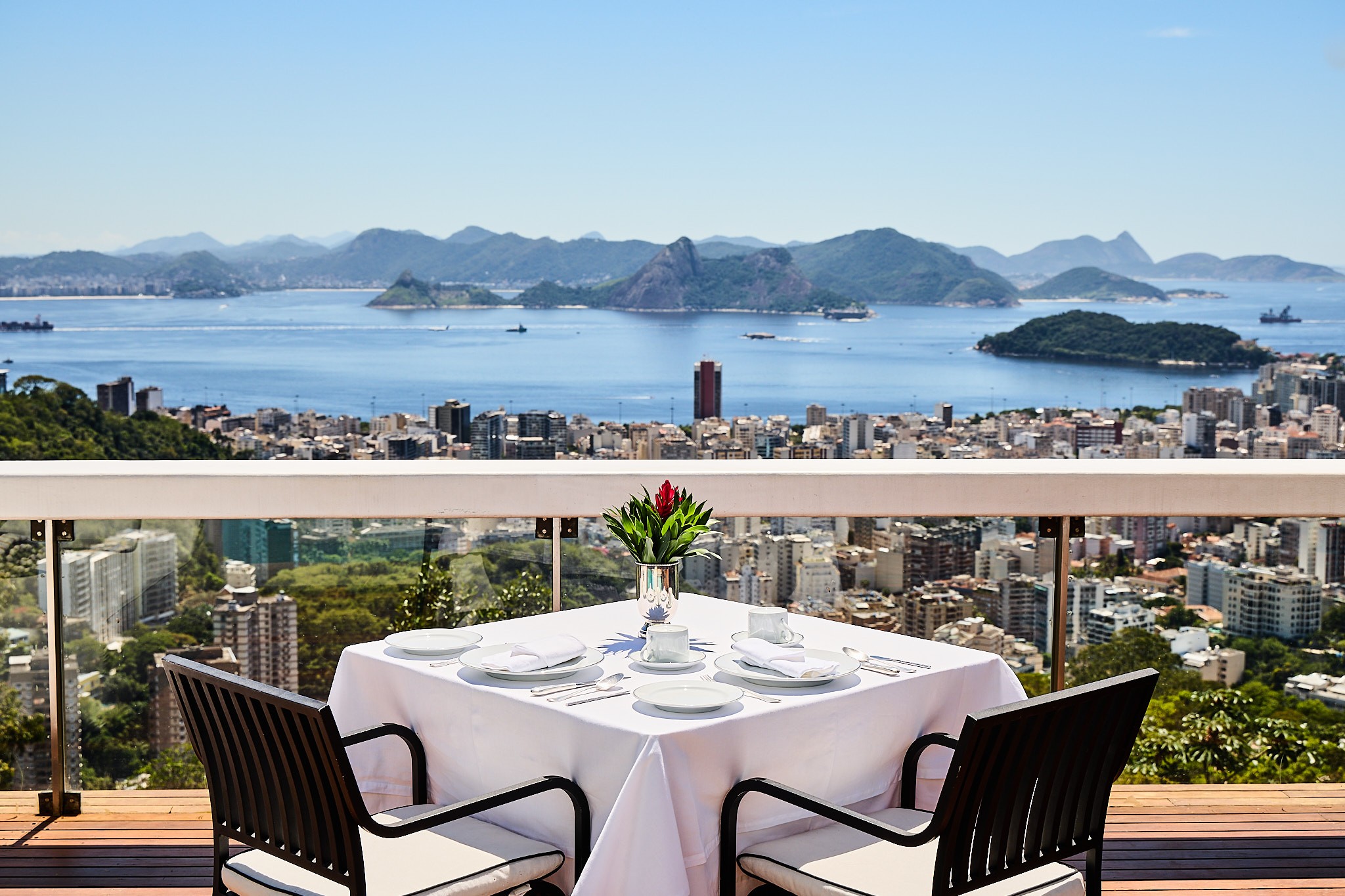 Mesa posta para duas pessoas em um terraço com vista panorâmica para a Baía de Guanabara, Rio de Janeiro. Pratos brancos, talheres e guardanapos sobre toalha branca, com um vaso de flores vermelhas no centro. Ao fundo, montanhas, prédios da cidade e o mar azul com barcos sob céu claro