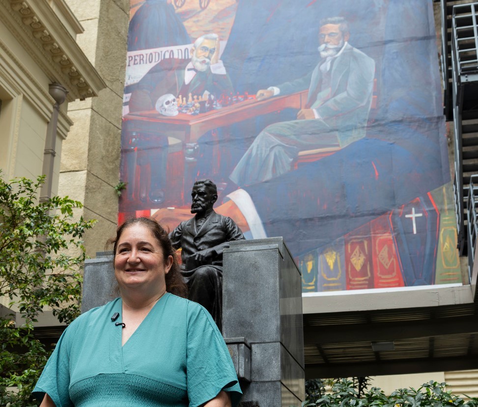 Mulher sorridente de blusa verde-água, com microfone preso à gola, em frente a uma estátua de bronze de um homem sentado e um mural colorido com figuras históricas e livros