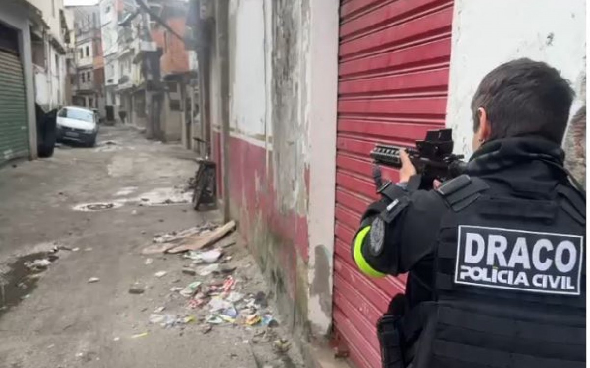 Agente da Polícia Civil com colete tático DRACO e fuzil, mirando em uma viela estreita e suja com lixo, carros e prédios antigos
