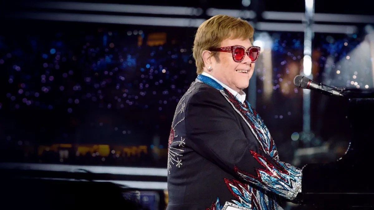 Elton John sorrindo, usando óculos de sol vermelhos e um paletó preto bordado com detalhes coloridos, tocando piano em um palco escuro com luzes azuis ao fundo