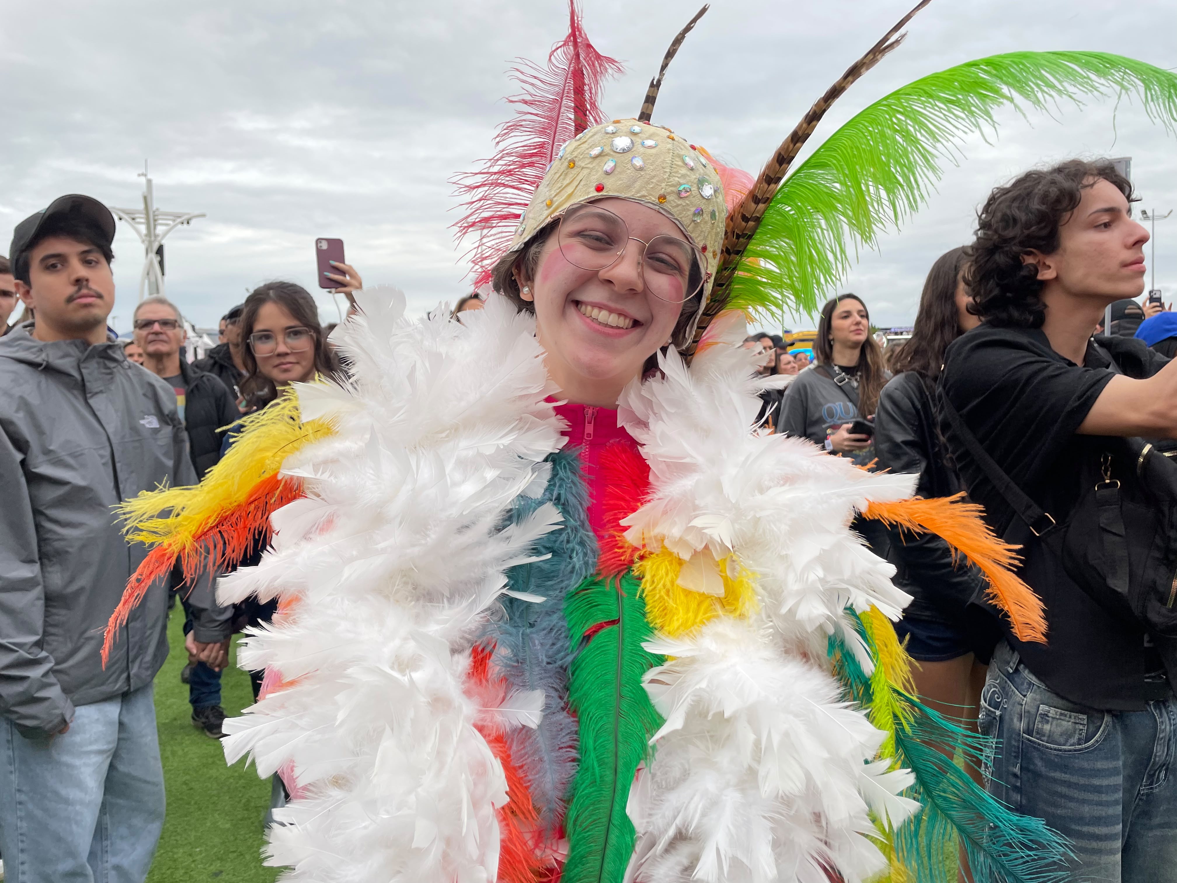 Jovem sorridente com óculos redondos, chapéu bege com pedras e penas coloridas, e um casaco de penas brancas, amarelas e laranjas, em meio a uma multidão em evento ao ar livre
