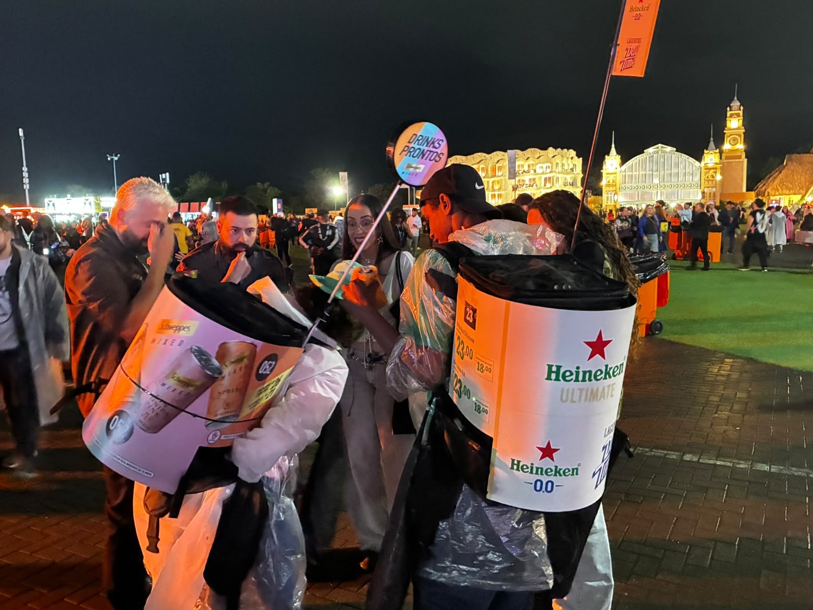 Pessoas em um evento noturno, com vendedores usando mochilas térmicas grandes. Um vendedor à esquerda tem uma mochila rosa com a marca Beefeater, e outro à direita tem uma mochila branca com a marca Heineken. Ao fundo, edifícios iluminados e uma multidão, sob um céu escuro