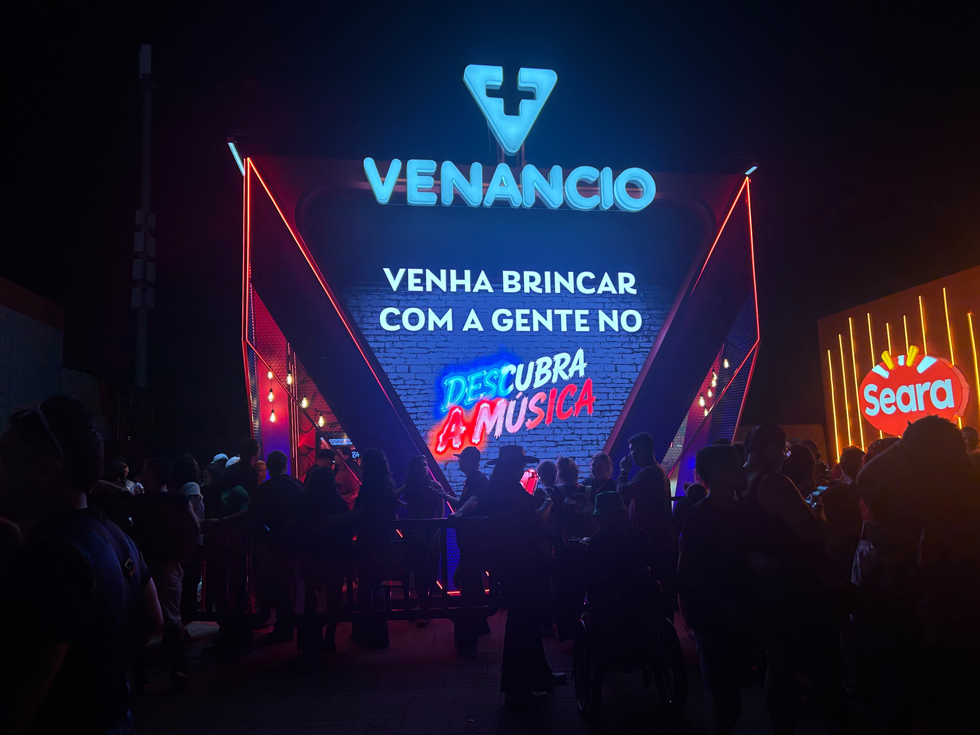 Pessoas aglomeradas em frente a um palco ou portal iluminado por luzes neon azuis e vermelhas. No centro, o letreiro VENANCIO com um símbolo de cruz, e abaixo, VENHA BRINCAR COM A GENTE NO DESCUBRA A MÚSICA. À direita, um painel laranja com o logo da Seara