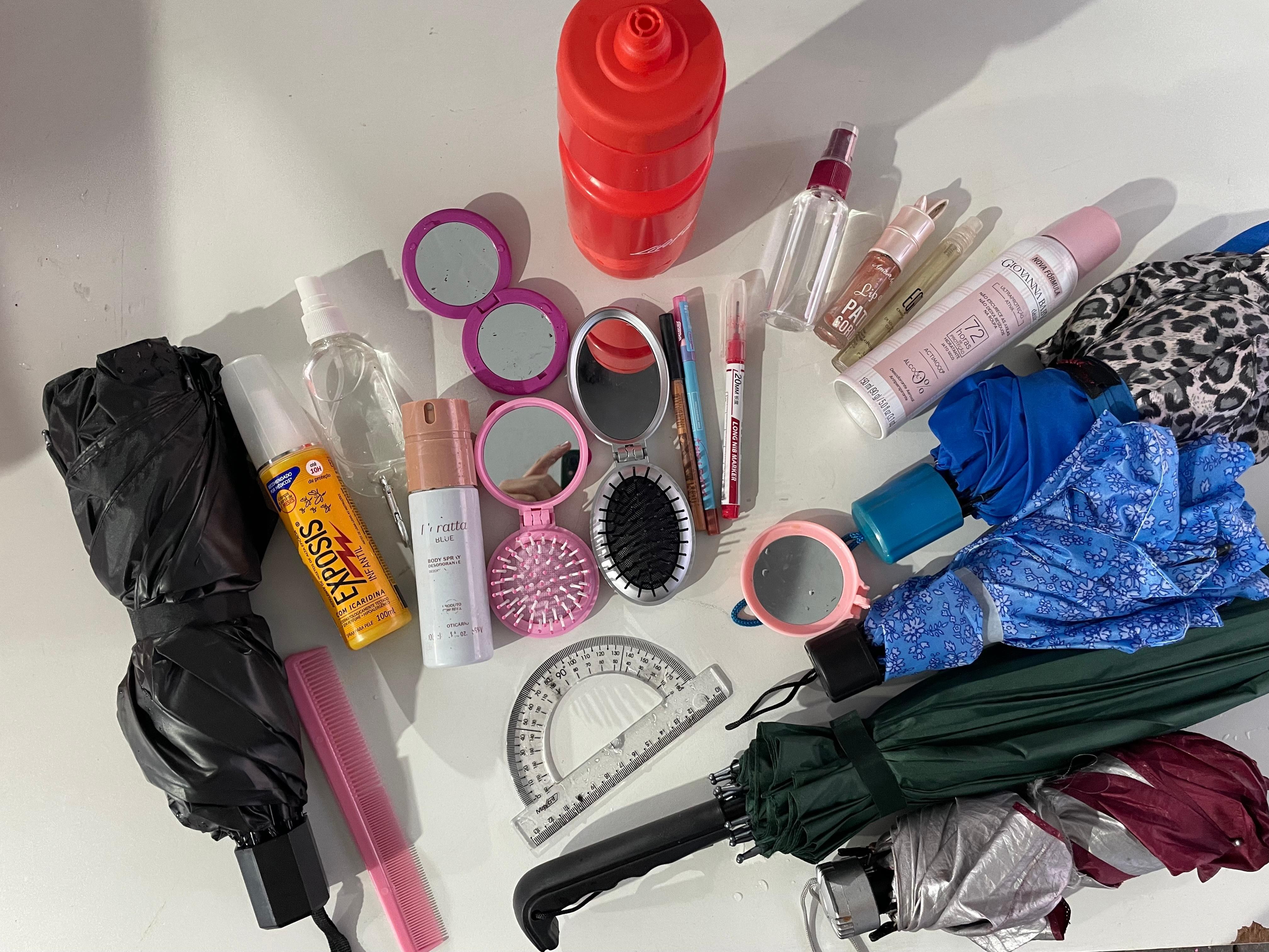 Diversos itens pessoais e de higiene espalhados sobre uma superfície branca, incluindo guarda-chuvas, espelhos compactos, escovas de cabelo, cosméticos, uma garrafa de água vermelha e um transferidor