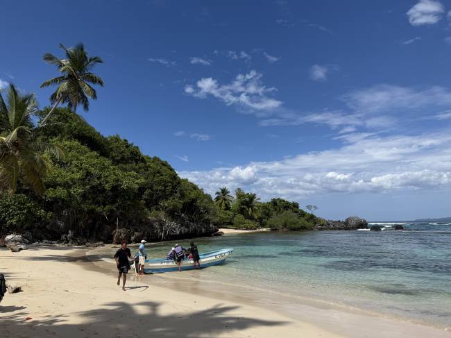 Pessoas desembarcando de um barco azul em uma praia de areia clara, com águas cristalinas e vegetação densa na encosta, sob céu azul com nuvens esparsas