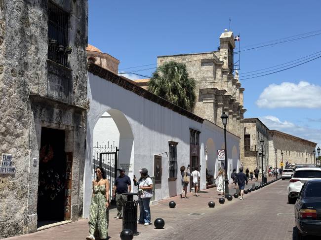 Rua histórica com edifícios de pedra e paredes brancas, sob céu azul. Pessoas caminham pela calçada de tijolos, com carros estacionados à direita. Uma mulher de vestido estampado verde está em primeiro plano, e ao fundo, uma igreja antiga com torre sineira e uma palmeira.