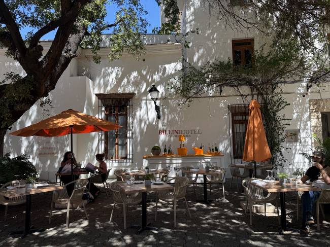 Pátio externo do Billini Hotel com mesas e cadeiras de vime, guarda-sóis laranja e um bar com garrafas. Duas mulheres conversam sob um guarda-sol e um homem bebe em outra mesa. Árvores frondosas sombreiam o chão de pedras, e a fachada branca do hotel tem janelas de madeira e ferro