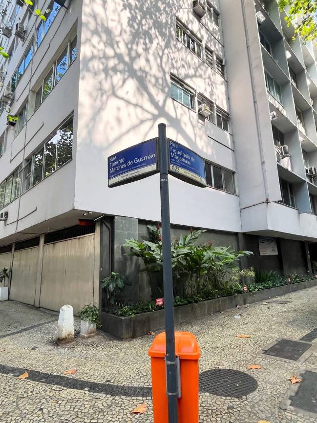 Placa de rua azul com Rua Tenente Marones de Gusm&atilde;o e Rua Figueiredo de Magalh&atilde;es 823, em poste cinza. Edif&iacute;cios claros com janelas refletindo o c&eacute;u, sombras de &aacute;rvores na parede. Cal&ccedil;ada de pedras portuguesas, lixeira laranja e folhagens verdes.