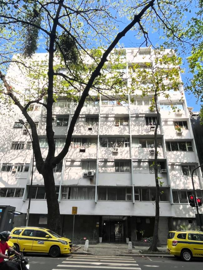 Edif&iacute;cio residencial branco de m&uacute;ltiplos andares com janelas e varandas, visto de baixo atrav&eacute;s de &aacute;rvores frondosas. Duas t&aacute;xis amarelos e uma faixa de pedestres na rua em primeiro plano.